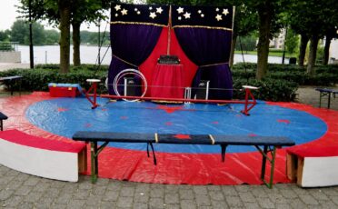 Circus podium met blauwe mat en rode rand, omringd door bomen en water.